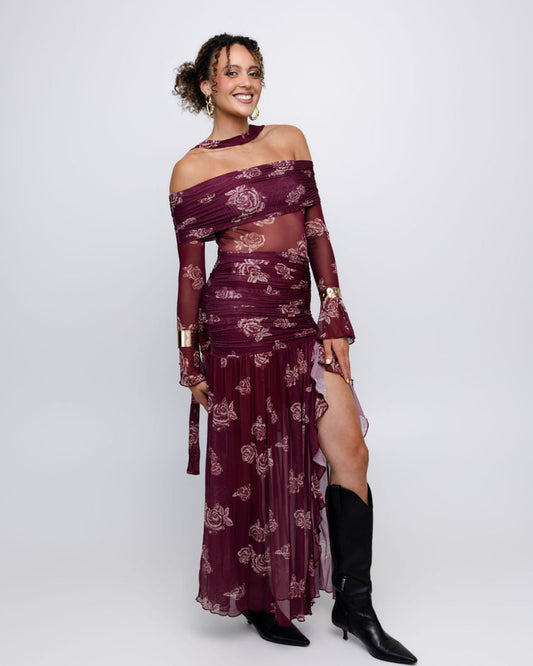 A woman models a long burgundy off-the-shoulder maxi dress with sheer sleeves and a matching pleated skirt, both decorated with a light floral pattern. She is wearing black pointed-toe heeled boots against a plain light gray studio background.