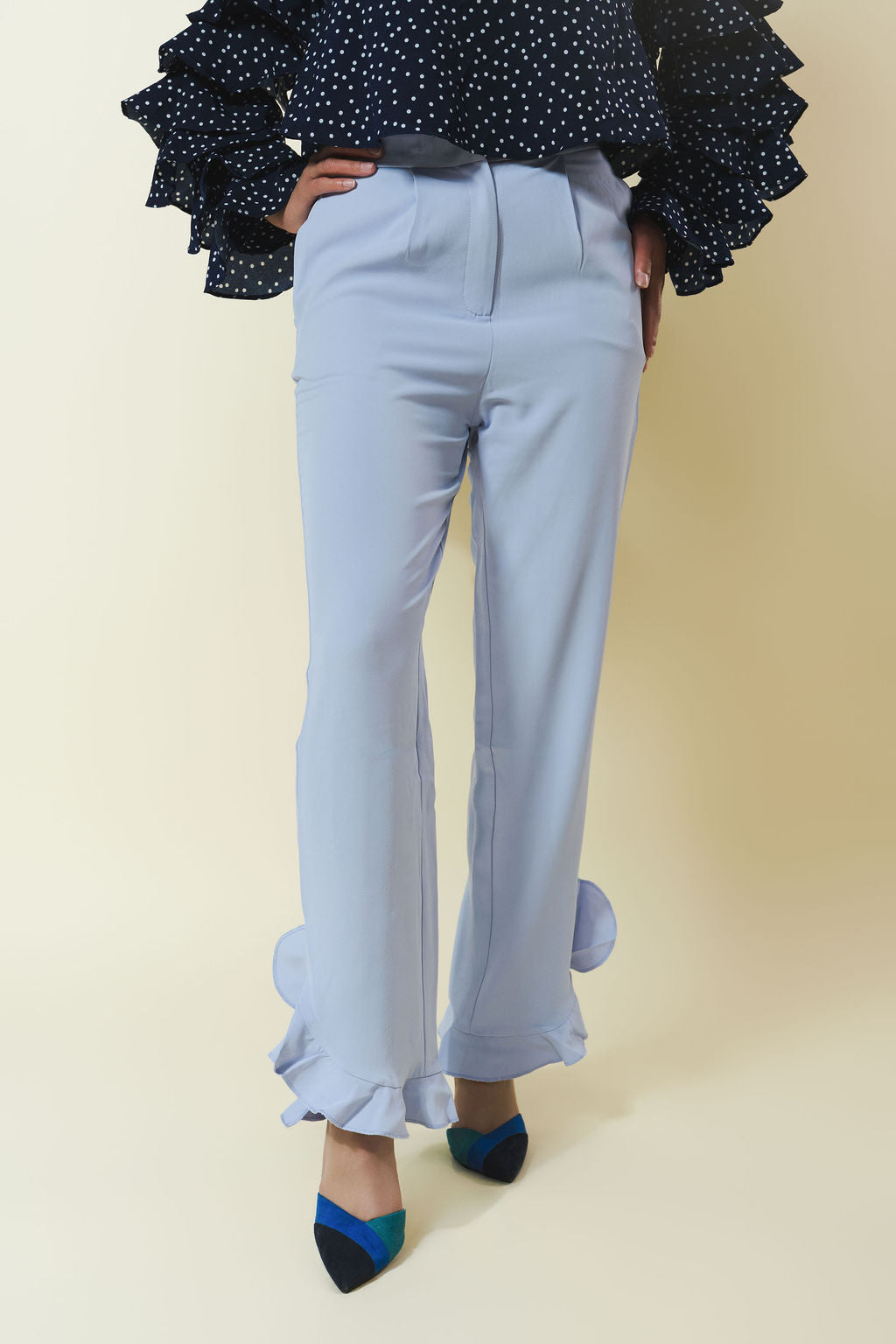 A person wearing light blue wide-leg trousers with ruffled hems, paired with a navy polka-dot blouse and blue-and-black pointed heels, against a plain beige studio background.
