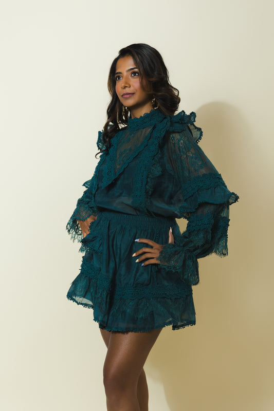 A woman modeling a dark teal green ruffled lace top and matching mini skirt against a plain beige studio background.