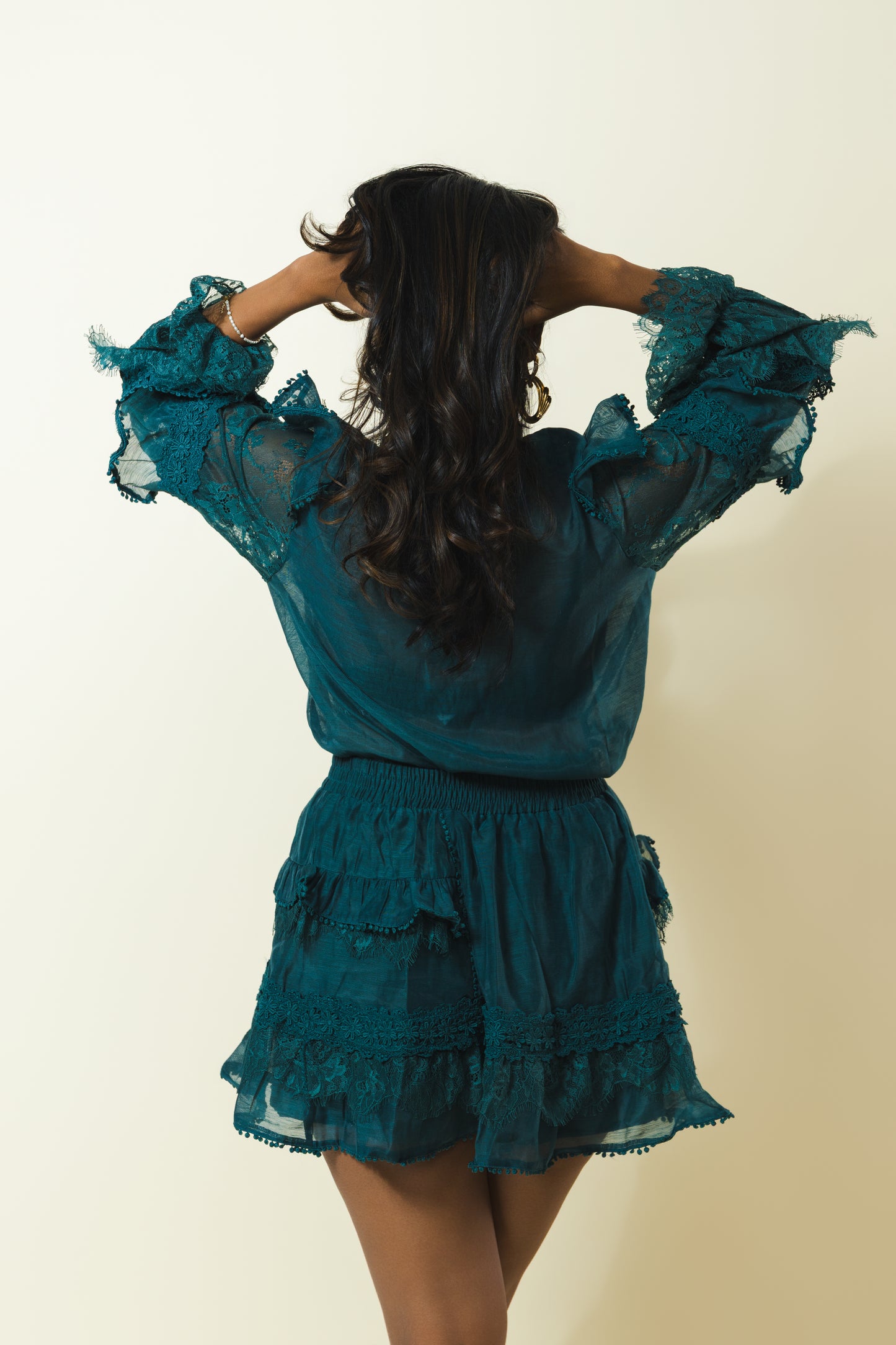 A woman viewed from behind wearing a teal blue sheer lace top and matching tiered skirt against a plain cream background.