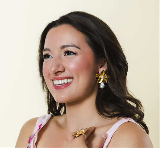 A smiling woman with dark hair models a sleeveless pink floral dress with vibrant magenta and red flowers, accessorized with gold jewelry against a solid cream background.