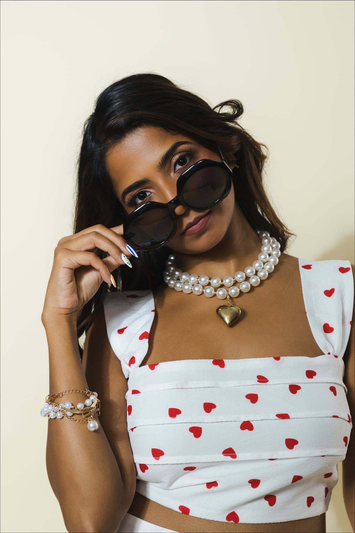 A woman wearing a white crop top with a red heart pattern, accessorized with a pearl necklace, a gold heart pendant, and pearl bracelets, holding a pair of black sunglasses against a light beige background.