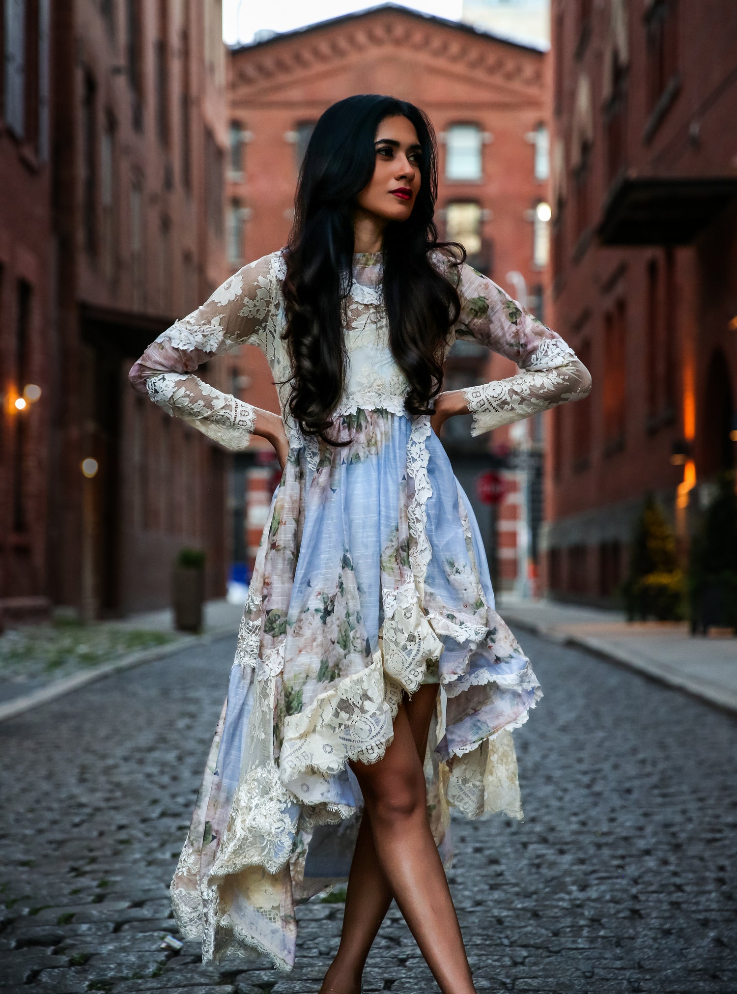 Woman in a floral dress standing on a cobblestone street with brick buildings.