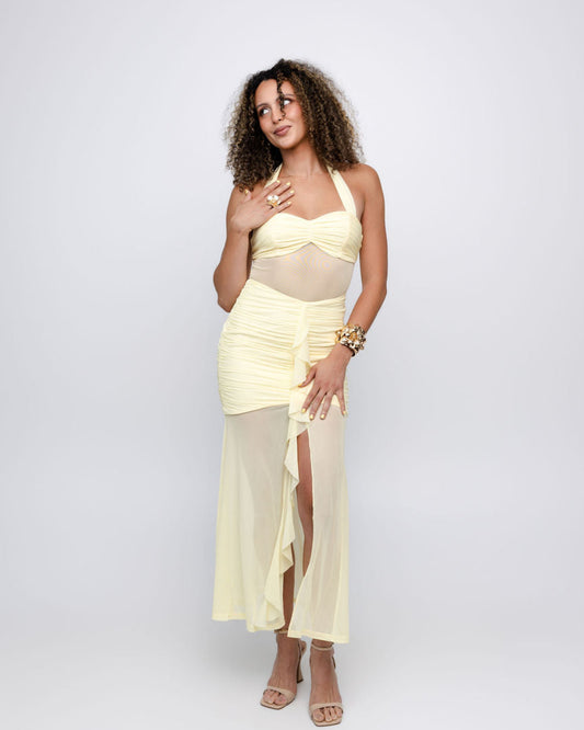 A woman modeling a pale yellow two-piece dress with a ruched halter top and a matching wrap skirt featuring sheer mesh panels and a thigh-high slit, posed against a light gray studio background.