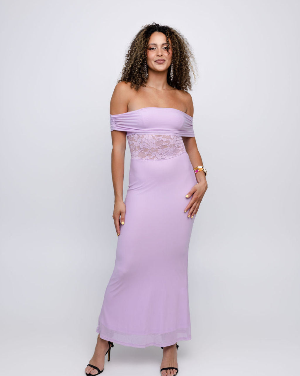 A woman modeling a lavender off-the-shoulder maxi dress with a sheer lace waist panel, photographed against a light gray studio background.