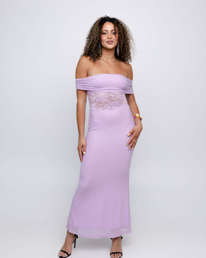 A woman modeling a lavender off-the-shoulder maxi dress with a sheer lace waist panel, photographed against a light gray studio background.