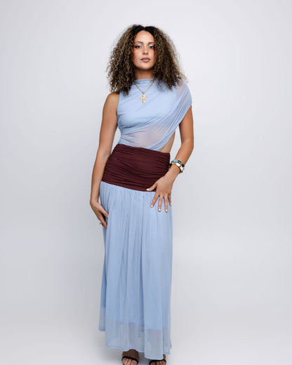 A woman modeling a light blue draped maxi dress with a sheer mesh overlay and a contrasting dark burgundy ruched waistband, set against a plain light gray studio background.