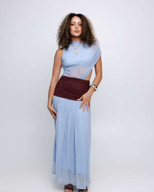 A woman modeling a light blue draped maxi dress with a sheer mesh overlay and a contrasting dark burgundy ruched waistband, set against a plain light gray studio background.