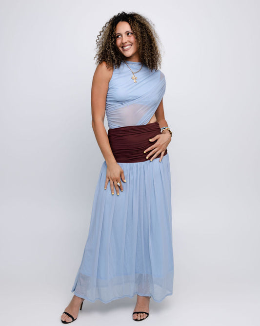 A woman modeling a sleeveless light blue draped maxi dress with a sheer mesh overlay and a wide dark burgundy wrap-style waist sash, photographed against a seamless light gray studio background.