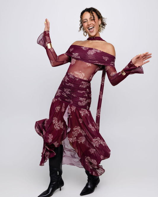 A woman modeling a sheer burgundy off-the-shoulder long-sleeve dress with a matching skirt and black pointed-toe heeled boots, posing against a light gray studio background.