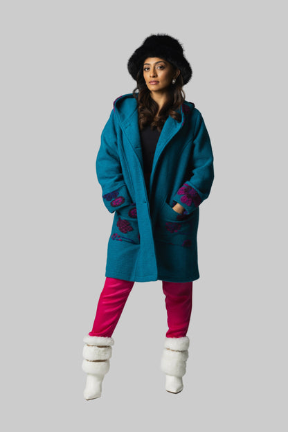 Woman wearing a teal coat with floral patterns, pink pants, and white boots on a gray background