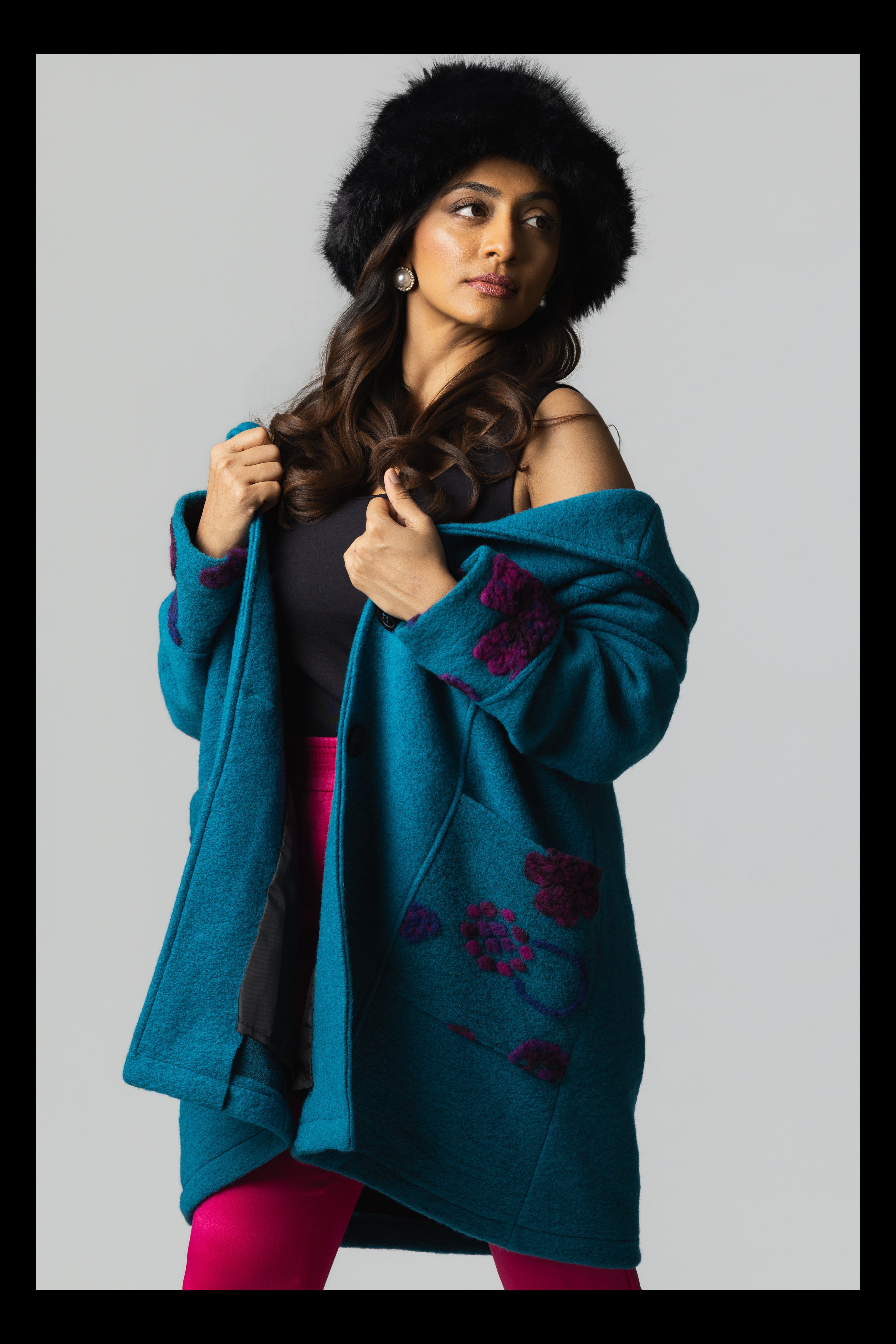 Woman wearing a teal coat with floral patterns and a black fur hat on a gray background