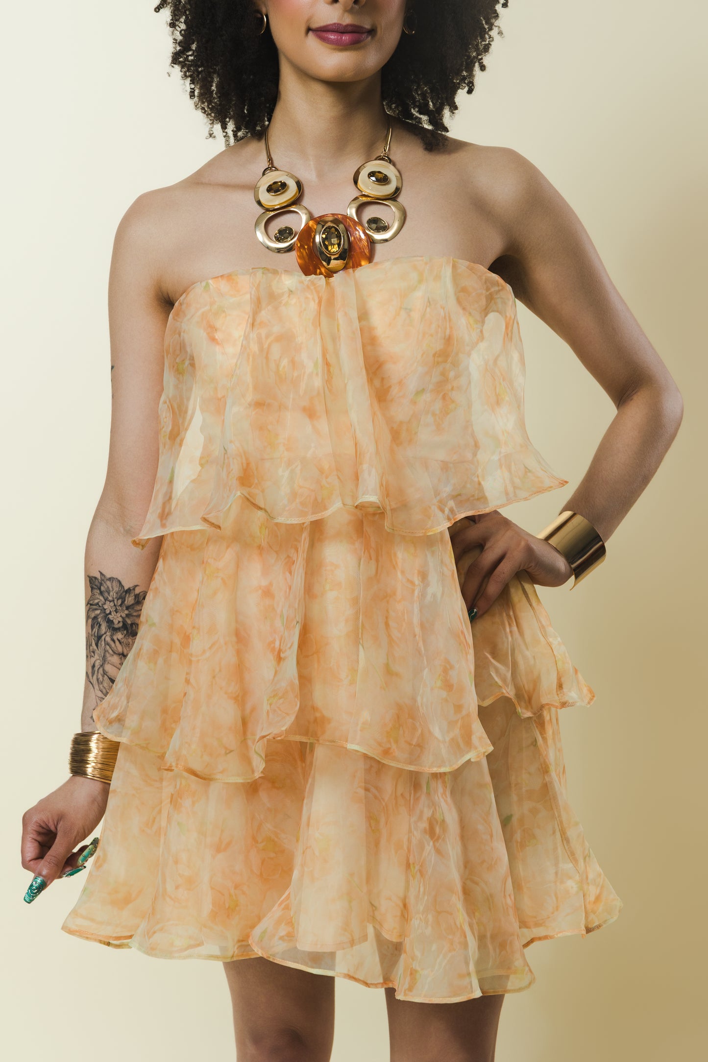 A woman wearing a strapless tiered mini dress in a light peach and orange floral print, styled with a chunky gold statement necklace and gold bracelets against a solid beige background.
