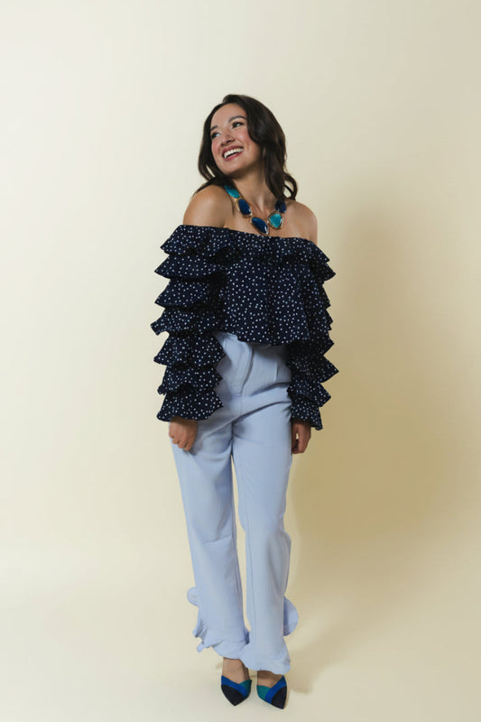 A woman modeling an off-the-shoulder navy blue top with white polka dots and layered ruffles, paired with light blue pants and blue heels, against a solid cream background.