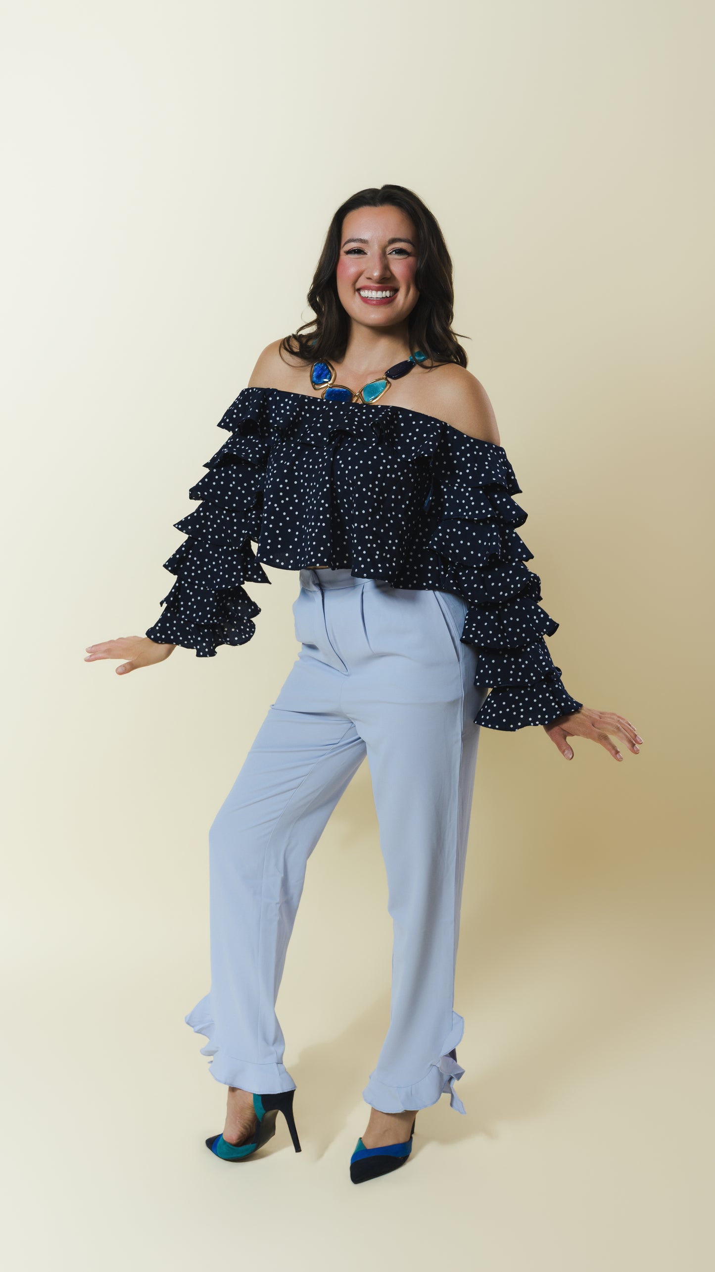 A woman modeling a dark navy off-the-shoulder ruffled top with white polka dots, paired with light blue high-waisted wide-leg pants and dark blue heels, against a solid cream background.