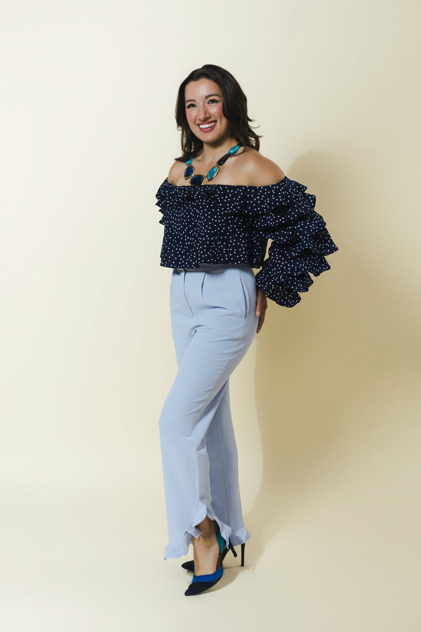 A woman modeling an off-the-shoulder navy blue top with white polka dots and ruffled sleeves, paired with light blue high-waisted wide-leg pants and black heels with blue accents, set against a solid cream background.