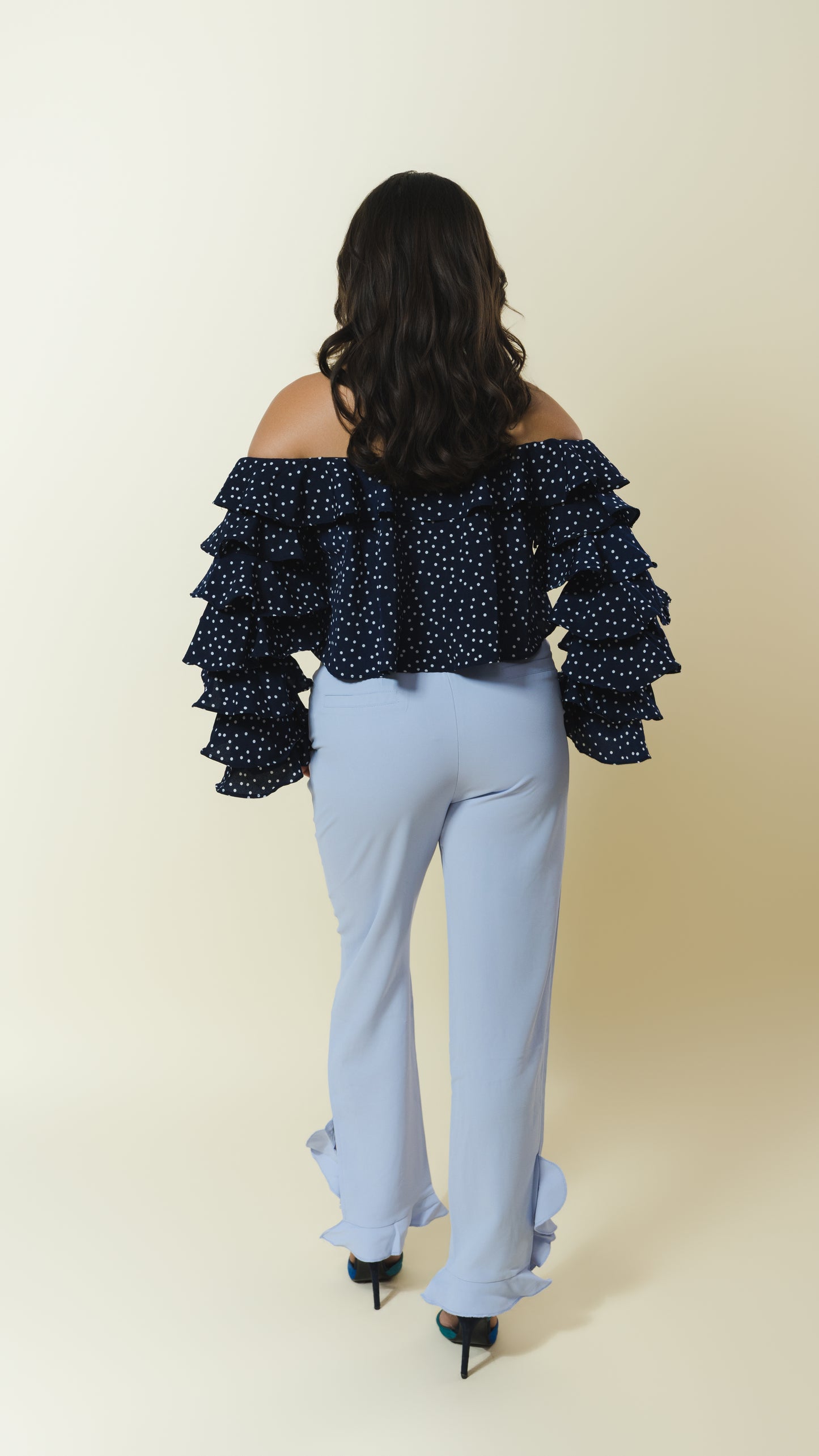 A woman viewed from the back wearing a navy blue off-the-shoulder ruffled top with white polka dots and light blue flared pants with ruffled hems, set against a plain beige studio background.