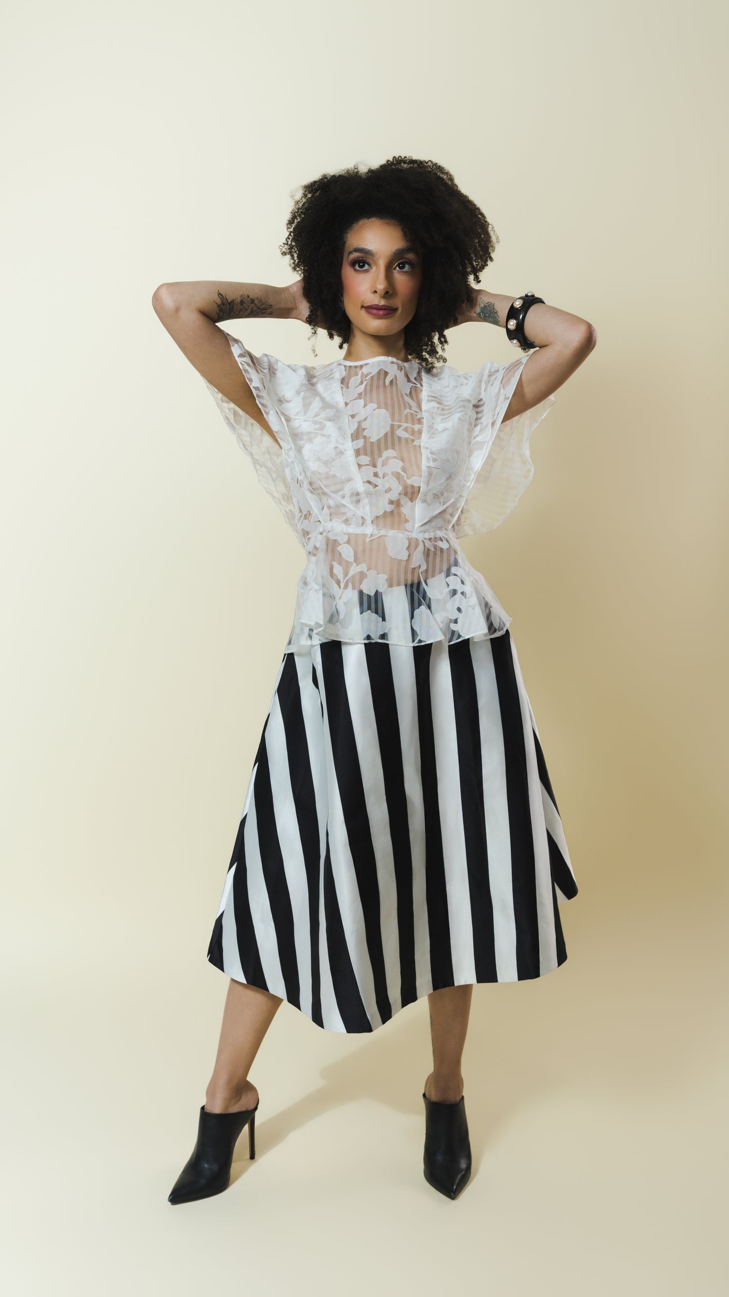 A model posing in a black and white striped midi skirt with a sheer white lace bodice and flutter sleeves, paired with black pointed-toe heels, against a solid beige background.