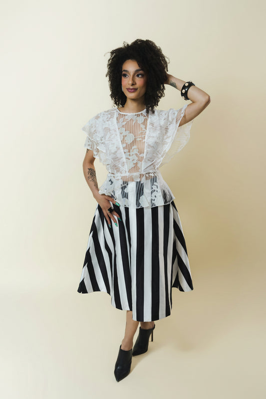 A model posing in a black and white striped midi skirt paired with a sheer white lace ruffled top and black pointed-toe heels, set against a solid cream background.