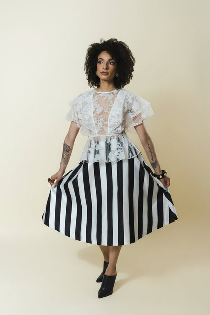 A model with curly dark hair is wearing a white lace short-sleeve top with a sheer V-neck and a black and white vertically striped skirt, holding out the skirt to show its volume. She is standing against a solid light beige background.