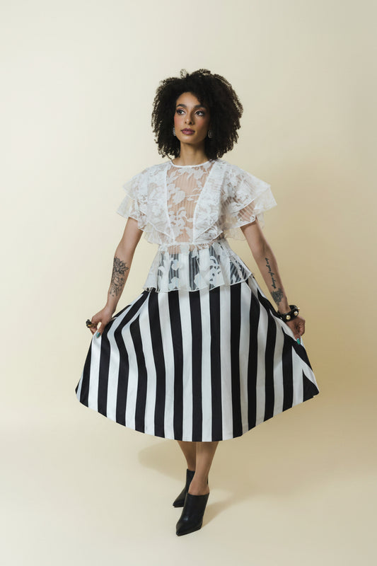 A model with curly dark hair is wearing a white lace short-sleeve top with a sheer V-neck and a black and white vertically striped skirt, holding out the skirt to show its volume. She is standing against a solid light beige background.