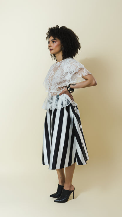 A woman stands in profile wearing a white lace short-sleeve top and a black-and-white vertically striped midi skirt, paired with black pointed-toe heels, against a plain warm beige studio background.