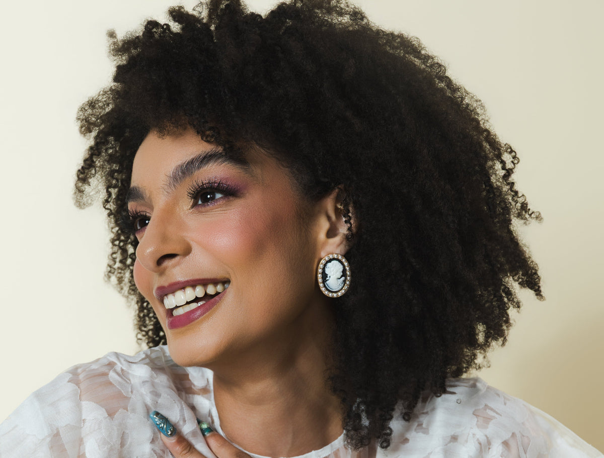 A woman with voluminous dark curly hair is shown from the chest up, wearing a sheer white floral-patterned ruffled blouse. She is smiling and looking to the side, with her hand resting on her chest.