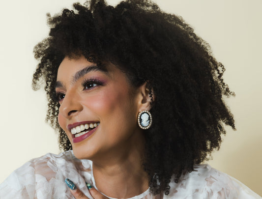 A woman with voluminous dark curly hair is shown from the chest up, wearing a sheer white floral-patterned ruffled blouse. She is smiling and looking to the side, with her hand resting on her chest.