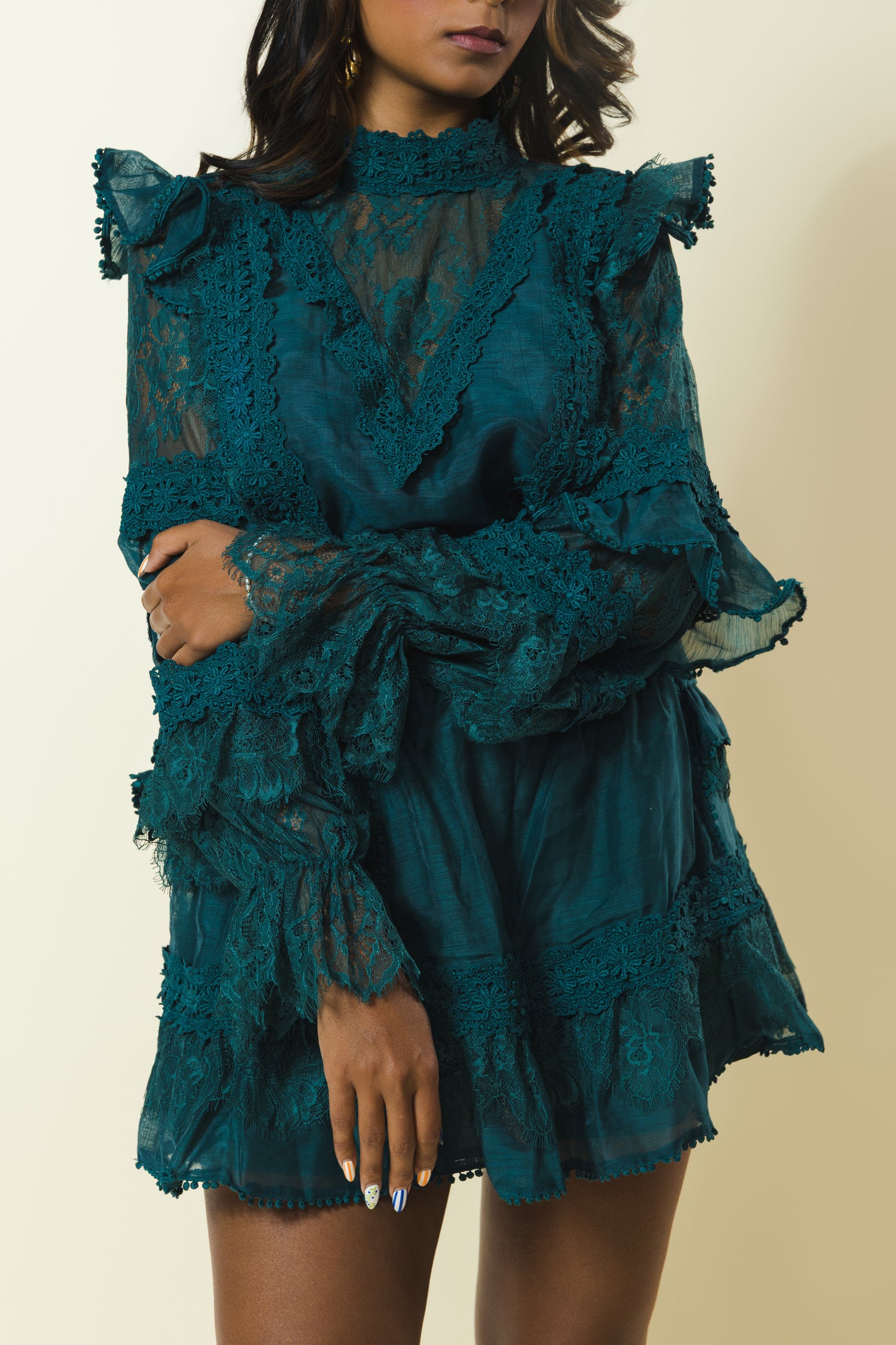 A model wearing a dark teal green ruffled lace mini dress with sheer long sleeves and a high neck, set against a plain beige background.