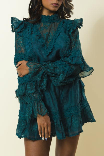 A model wearing a dark teal green ruffled lace mini dress with sheer long sleeves and a high neck, set against a plain beige background.