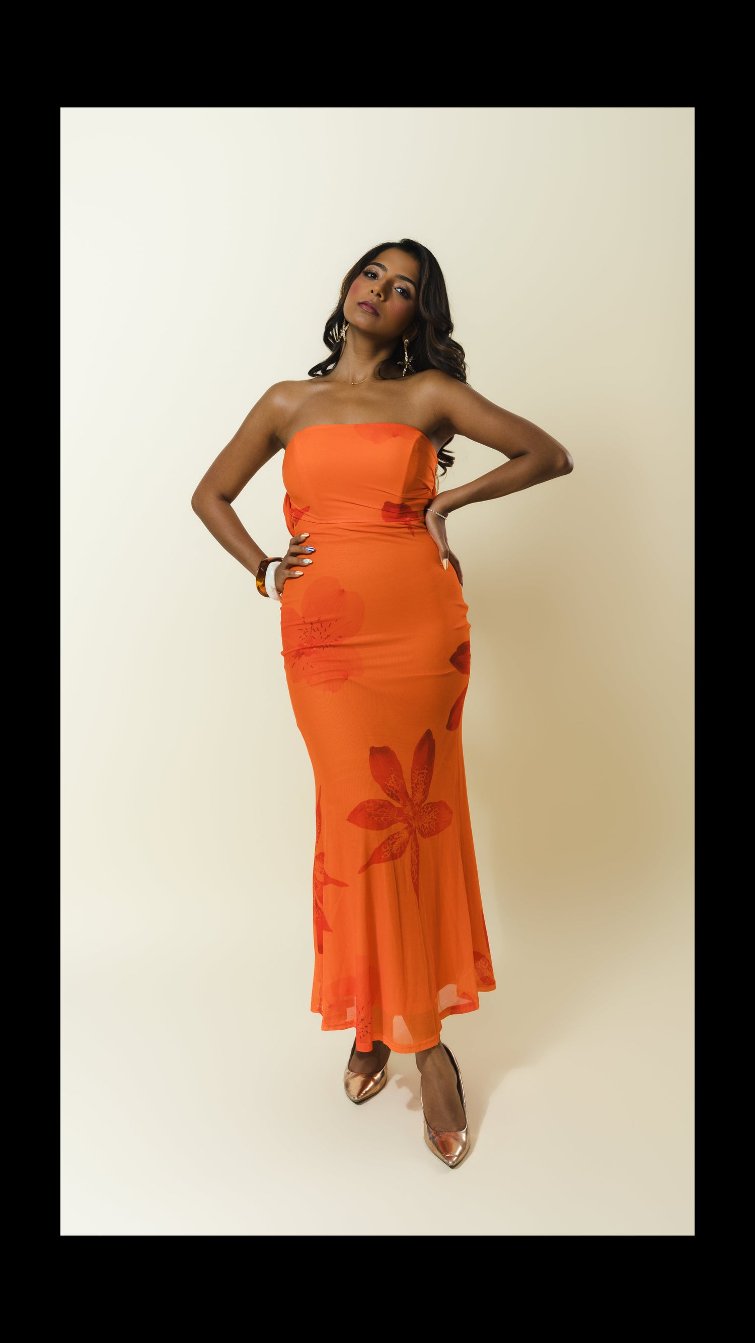 A woman modeling a strapless orange maxi dress with a tonal floral pattern, standing against a plain beige studio background.
