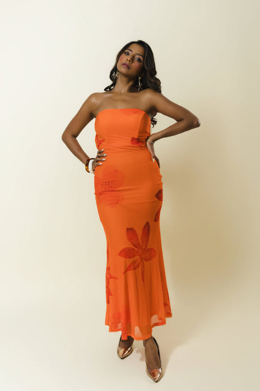 A woman modeling a strapless orange maxi dress with a tonal floral pattern, standing against a plain beige studio background.