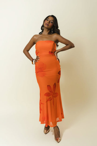 A woman modeling a strapless orange maxi dress with a tonal floral pattern, standing against a plain beige studio background.