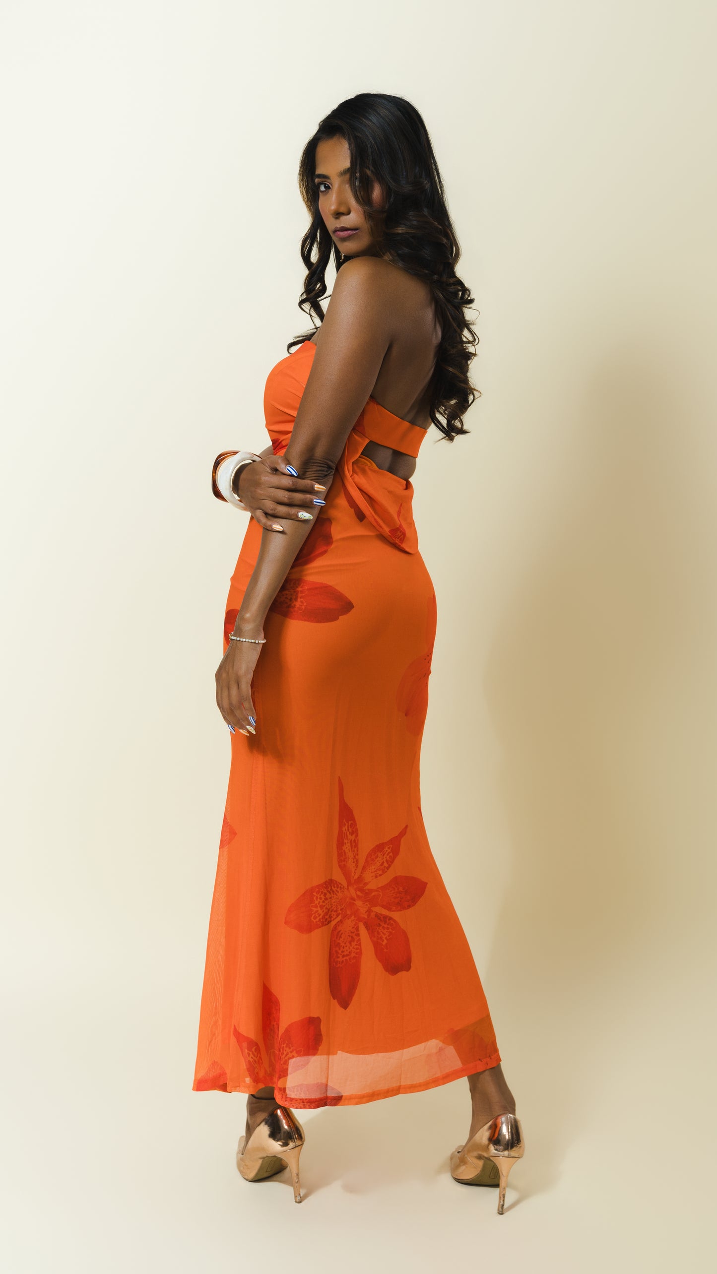 A woman modeling a bright orange sleeveless maxi dress with a floral pattern and a cutout at the waist, standing against a plain beige background.