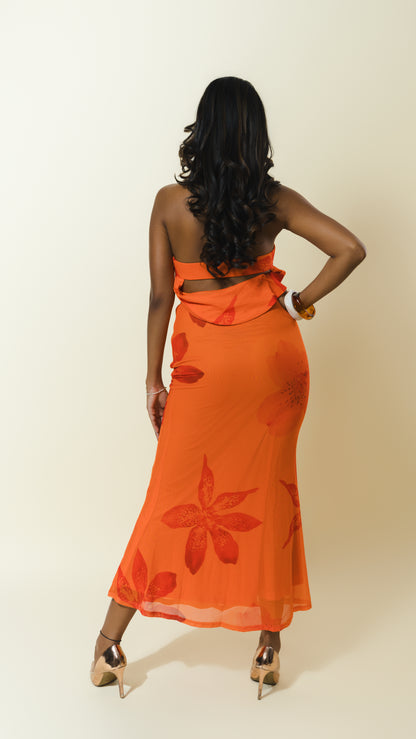 A woman shown from the back wearing a bright orange strapless maxi dress with a large red floral pattern, paired with metallic gold high heels, against a plain beige studio background.