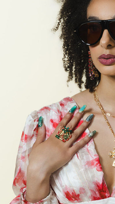 A woman with curly black hair wearing a white and red tie-dye long-sleeve dress, gold cross necklace, and sunglasses, posing against a solid beige background.