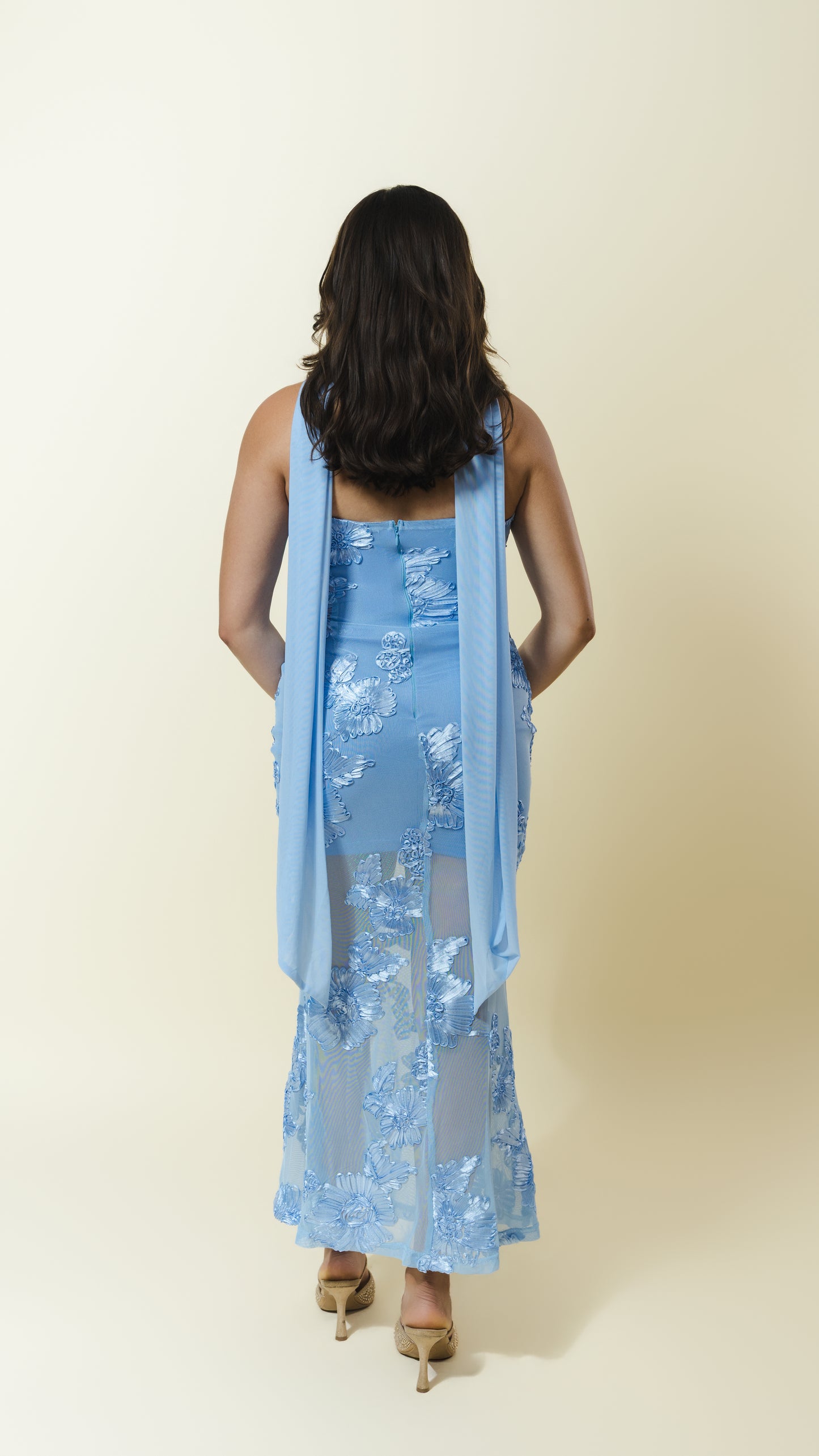 A woman viewed from behind wearing a light blue sleeveless dress with floral lace applique and a matching sheer draped overlay, paired with tan high heels, against a plain cream studio background.