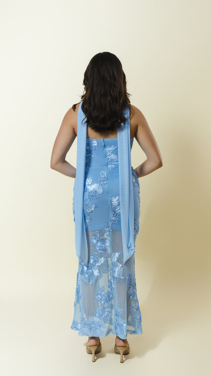 A woman viewed from behind wearing a light blue sleeveless dress with a matching sheer draped scarf, standing against a plain cream background.