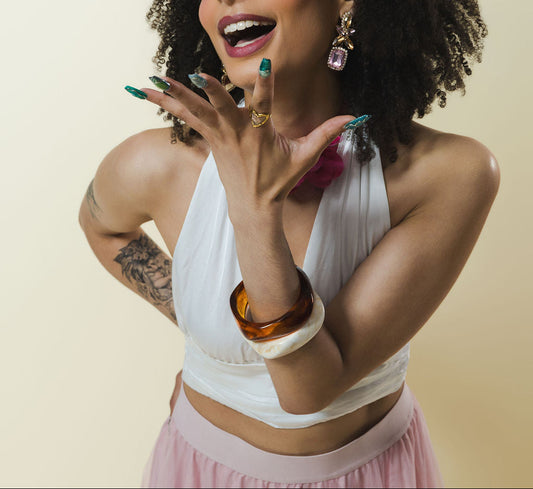 A woman with voluminous dark curly hair, wearing a white halter crop top and a light pink skirt, posing against a solid beige background. She is looking upward and to the side with an expressive, joyful face, holding her hands near her face.