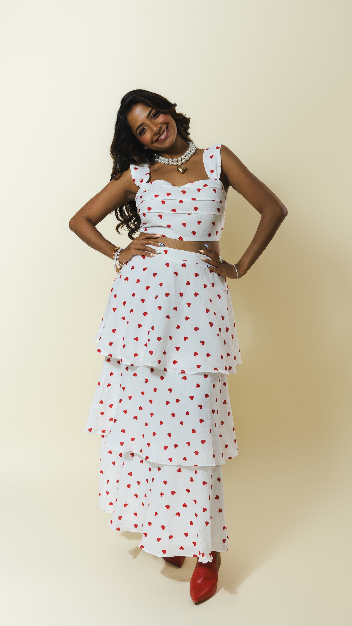 A woman modeling a white sleeveless tiered maxi dress with a red heart pattern, styled with a matching crop top, layered skirt, and red pointed shoes against a plain beige studio background.