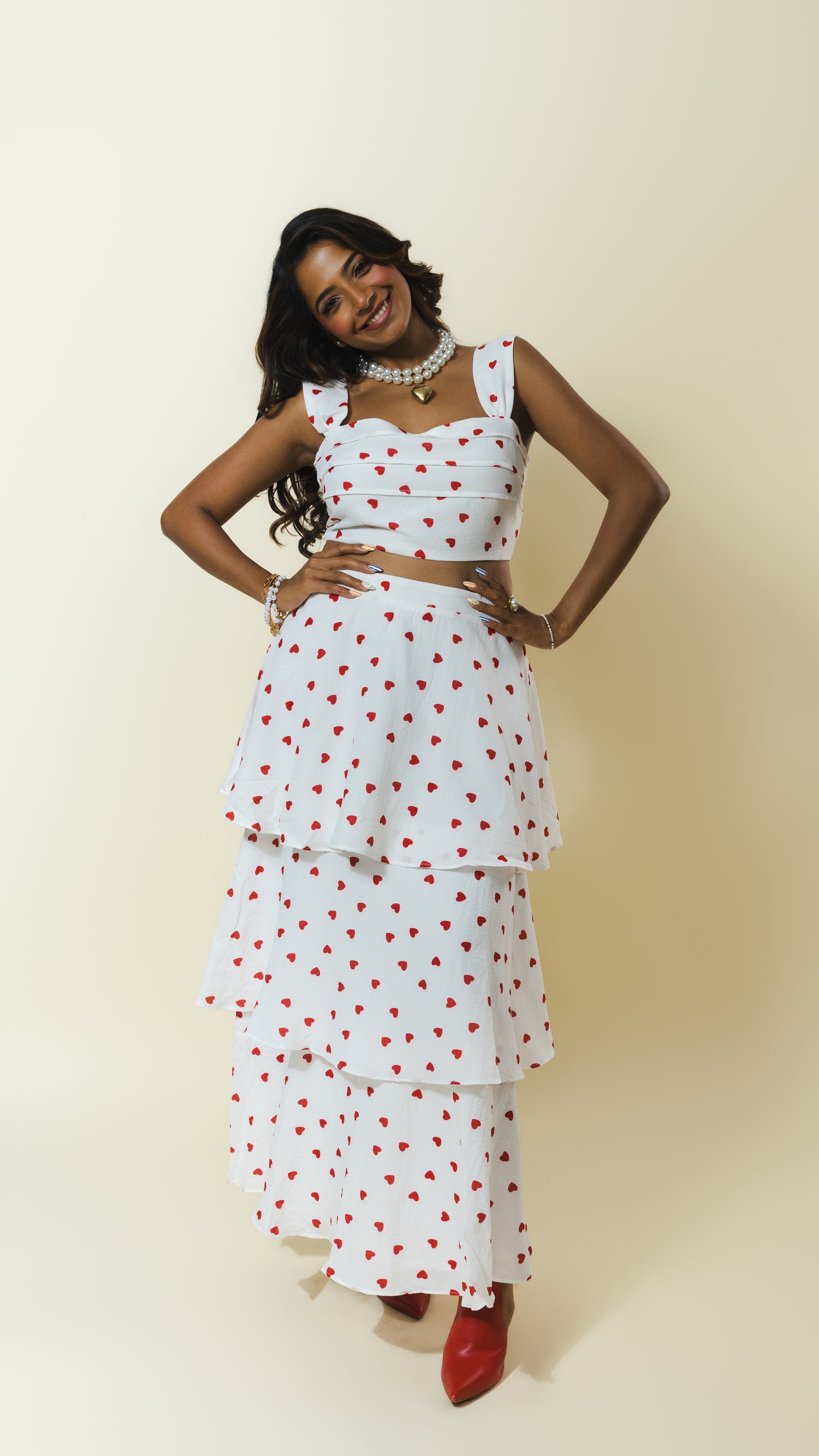 A woman modeling a white sleeveless tiered maxi dress with a red heart pattern, styled with a matching crop top, layered skirt, and red pointed shoes against a plain beige studio background.