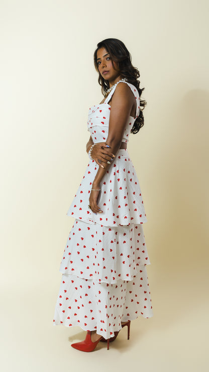 A woman modeling a sleeveless white tiered maxi dress with a red heart pattern, paired with red pointed-toe heels, set against a plain beige studio background.