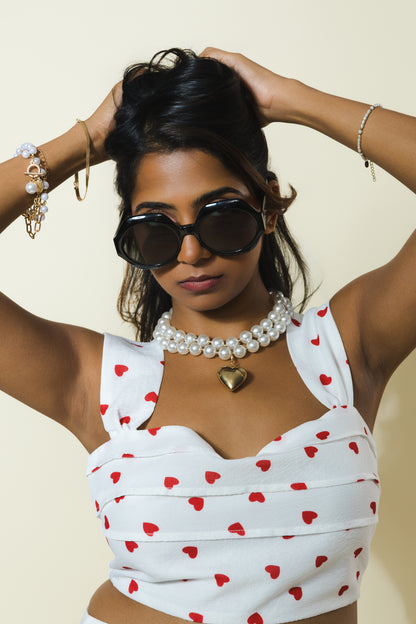 A woman wearing oversized black sunglasses, a pearl collar necklace with a gold heart pendant, and a white crop top with red hearts, posing against a light beige background.