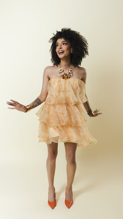 A woman modeling a strapless ruffled mini dress in a light peach and orange tie-dye pattern, paired with orange pointed-toe heels and a chunky gold statement necklace, set against a solid cream background.